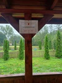 słup altany i część zadaszenia od środka z tabliczką informującą o współfinansowaniu projektu, w tle trawa, krzewy