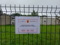 ogrodzenie panelowe, na nim tabliczka z informacją o dofinansowaniu projektu, za ogrodzeniem trawa, dalej budynek gospodarczy