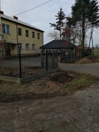 ogrodzenie panelowe, za nim utwardzony plac, budynek, altana, za nią drzewa