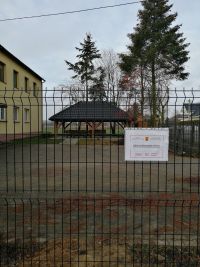 ogrodzenie panelowe, na nim tabliczka z informacją o dofinansowaniu. W tle utwardzony plac, budynek, altana, za nią drzewa